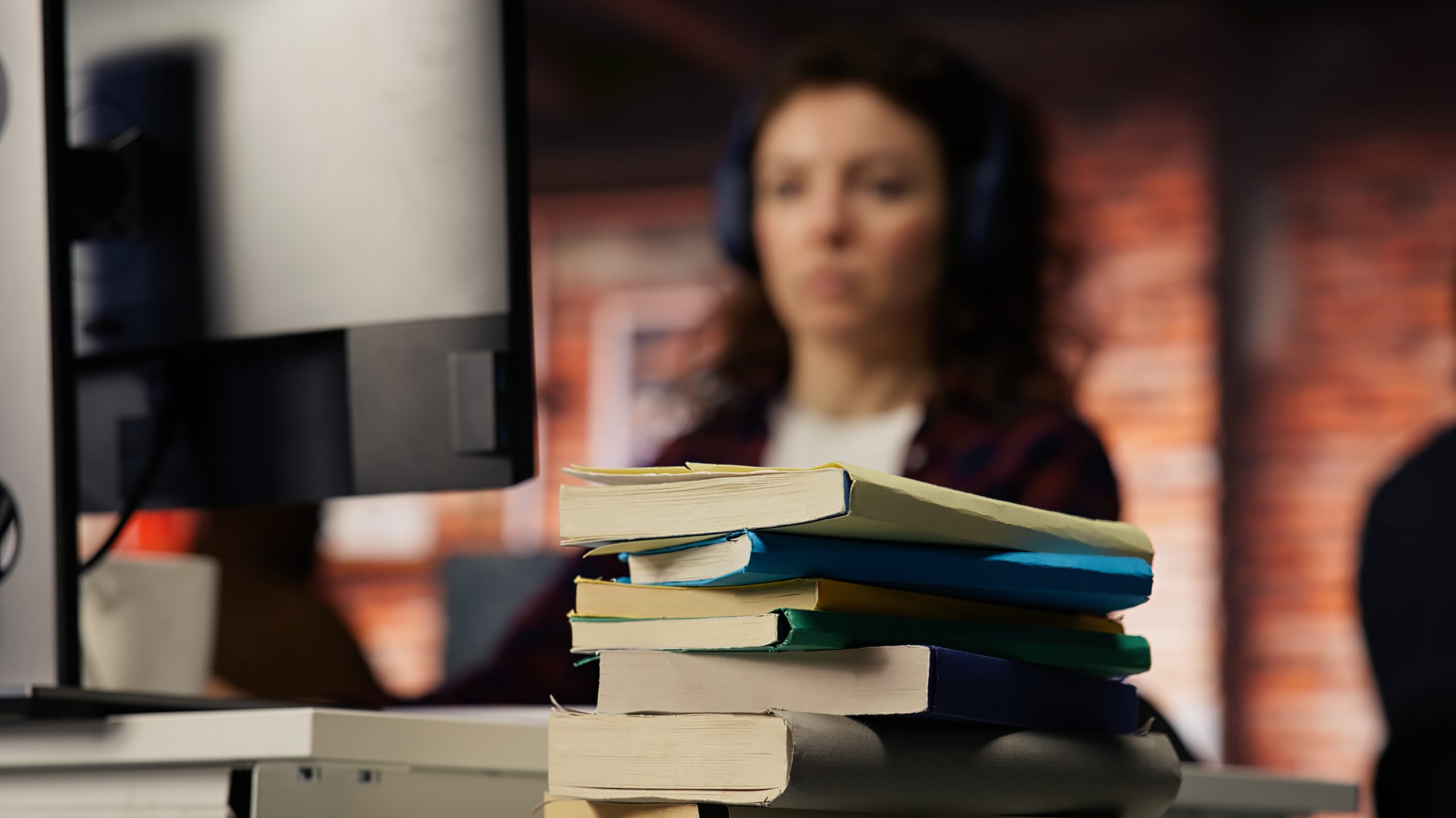 Focus on stack of books on startup office desk with woman in blurred background typing on PC, solving tasks. Pile of books in venture company workspace in front of employee working, camera B closeup